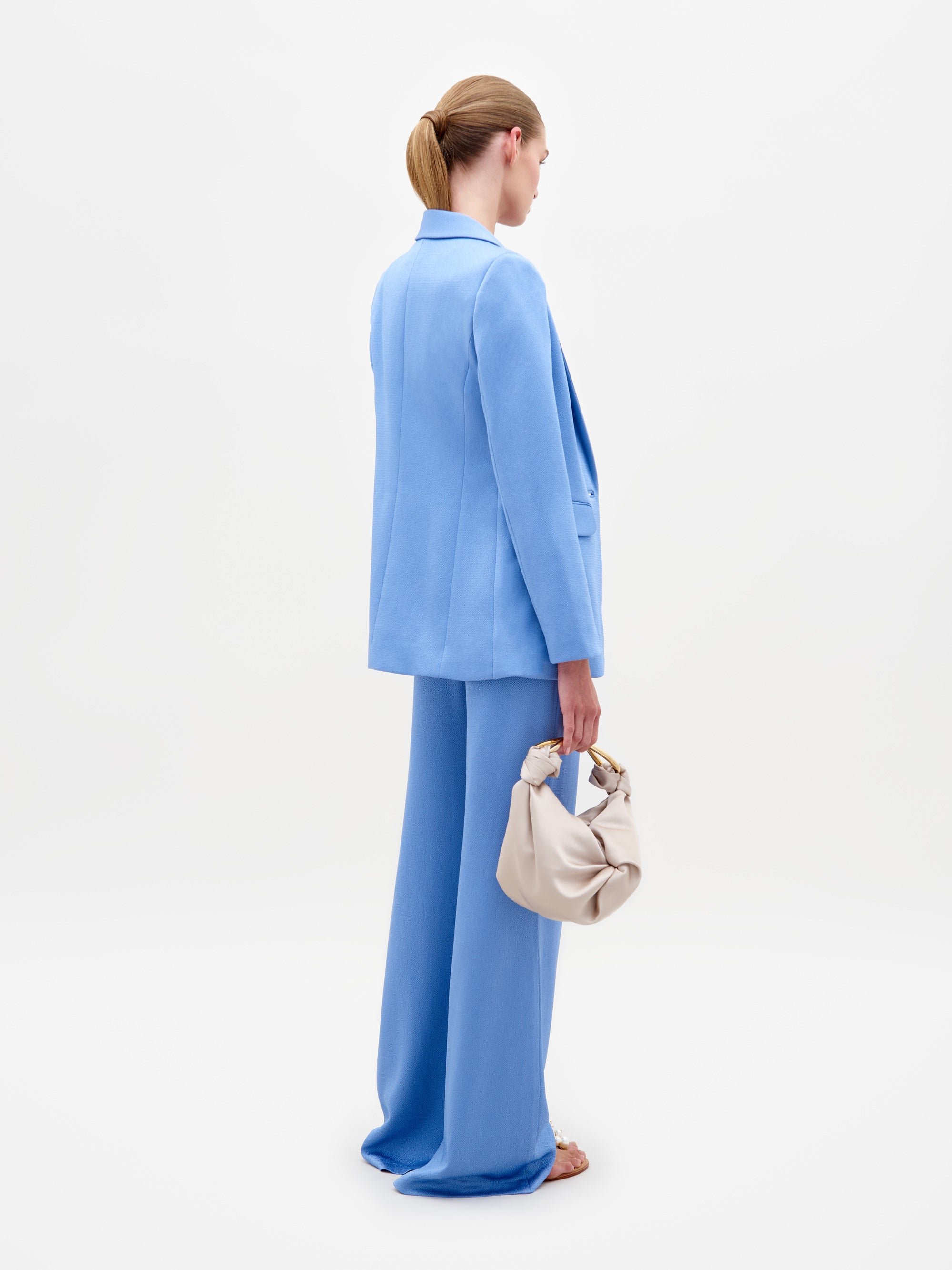 A woman in a light blue Rebeca Jacket with hammered satin, single-button closure, and matching knotted top and pants holds sunglasses against a plain white background.