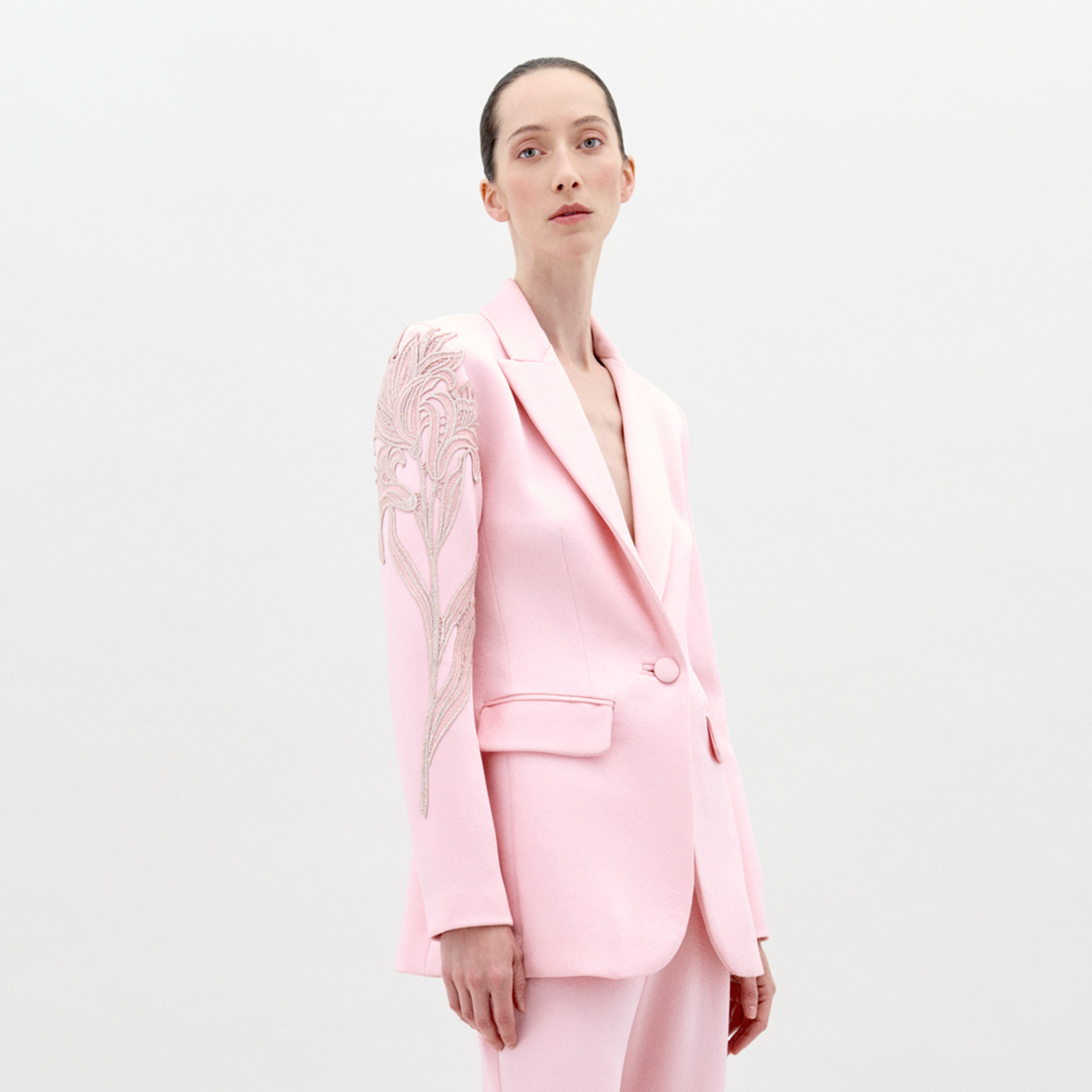 Profile view of a person with long hair tied back, wearing the Rebeca Jacket Pink, a light pink blazer adorned with intricate embroidery on the shoulder; pre-order now for delivery by February 1st, 2025.