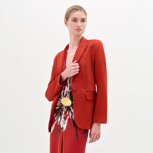 A woman with blonde hair tied back wears the Rebeca Jacket, a red fitted blazer, over a patterned red dress with a gold circular belt. She stands against a plain white background, looking slightly to the side.
