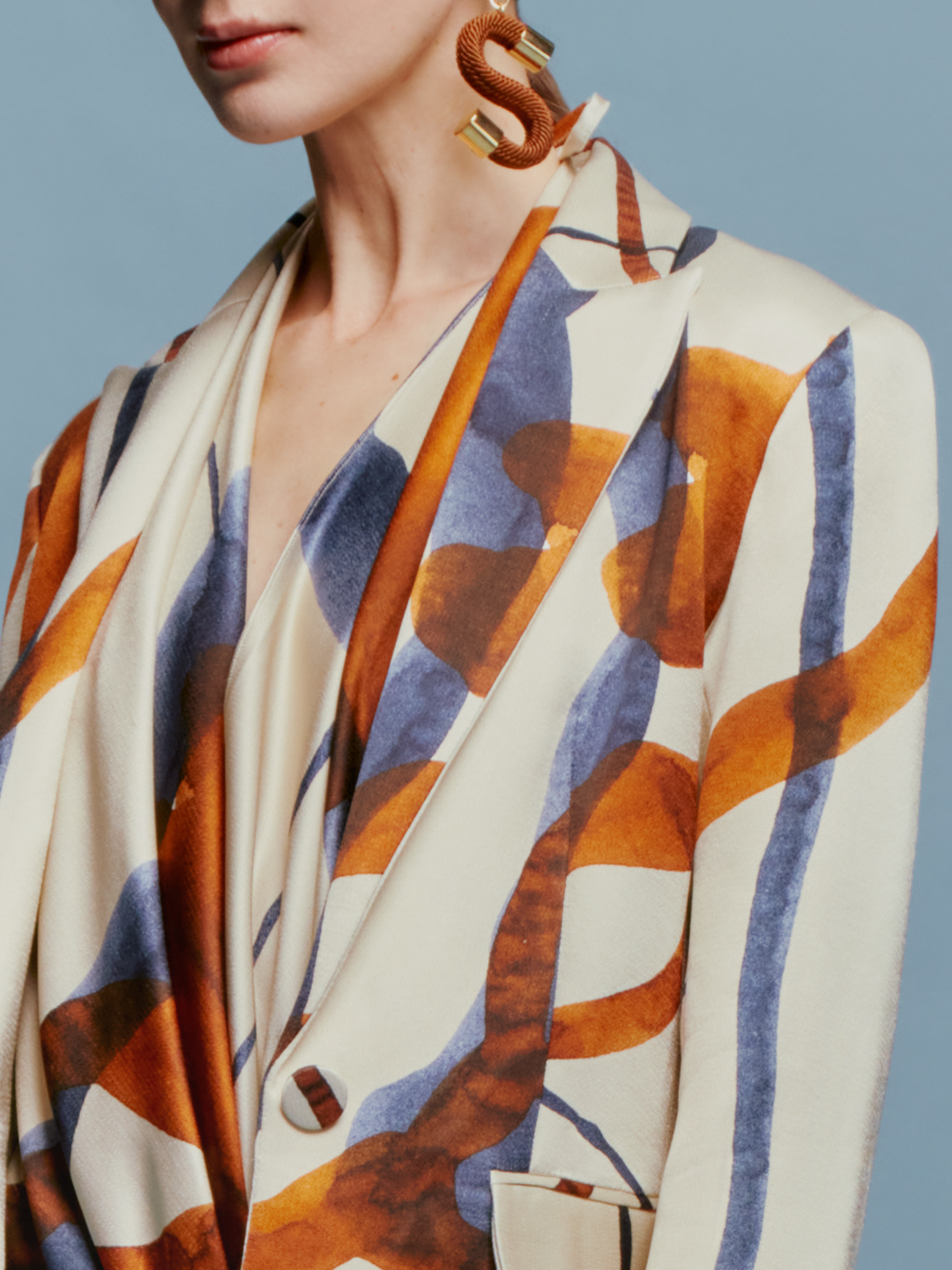 A woman with tied back hair stands against a blue background, wearing the Rebeca Jacket in Cobalt Copper Abstract with a matching dress, and large, abstract earrings, ready for her feature on September 2nd.