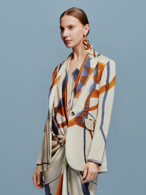 A woman with tied back hair stands against a blue background, wearing the Rebeca Jacket in Cobalt Copper Abstract with a matching dress, and large, abstract earrings, ready for her feature on September 2nd.