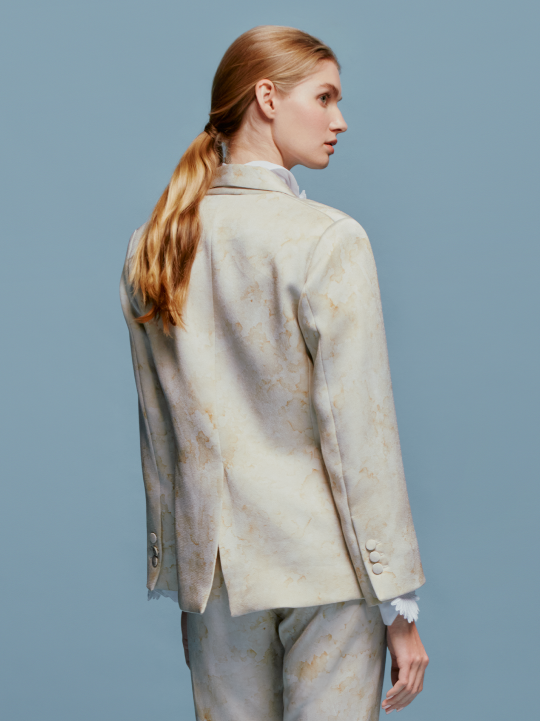 A person stands against a plain blue background wearing the Rebeca Jacket Cream Abstract Print draped over their shoulders, complemented by a white blouse with a ruffled collar, matching pants, and a gold belt with an "S" charm. The ensemble exudes timeless elegance, perfect for events like August 19th or September 2nd.