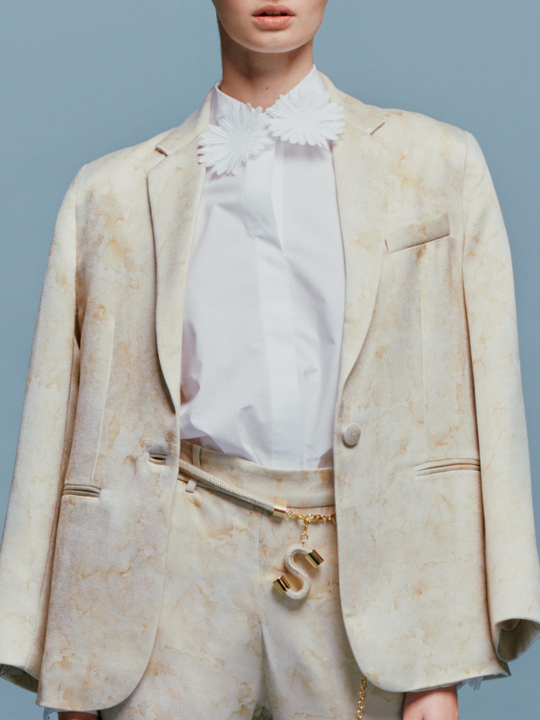 A person stands against a plain blue background wearing the Rebeca Jacket Cream Abstract Print draped over their shoulders, complemented by a white blouse with a ruffled collar, matching pants, and a gold belt with an "S" charm. The ensemble exudes timeless elegance, perfect for events like August 19th or September 2nd.