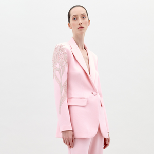 A person with slicked-back hair wears the Rebeca Jacket, a light pink hammered satin blazer with an embroidered floral embellishment on one shoulder, standing against a plain white background.