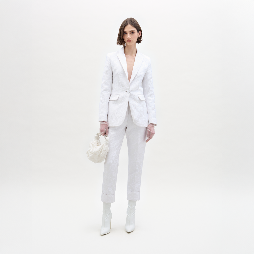 A person in a white Moad Pant suit and boots holds a textured handbag, standing against a plain white background.