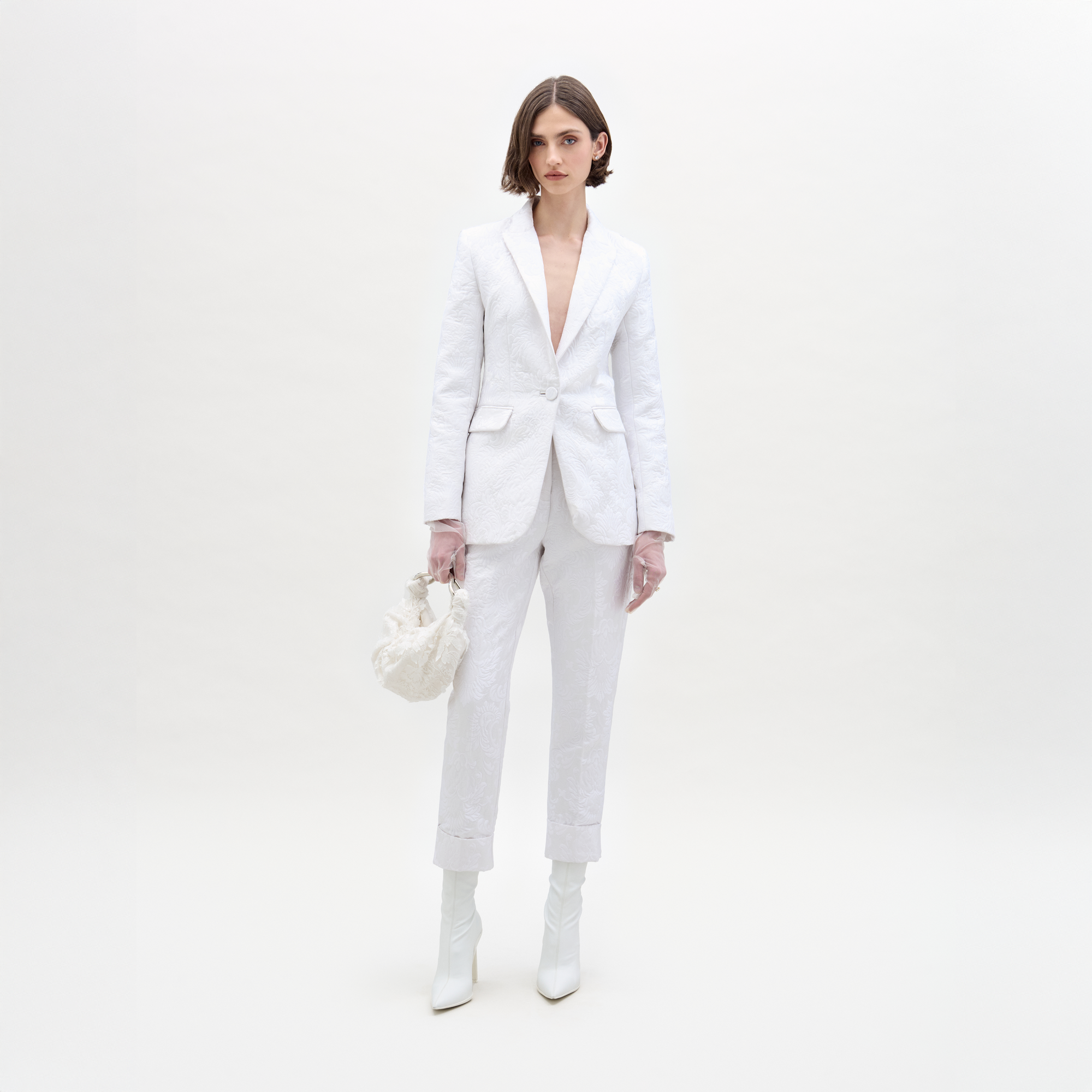 A person in a white Moad Pant suit and boots holds a textured handbag, standing against a plain white background.