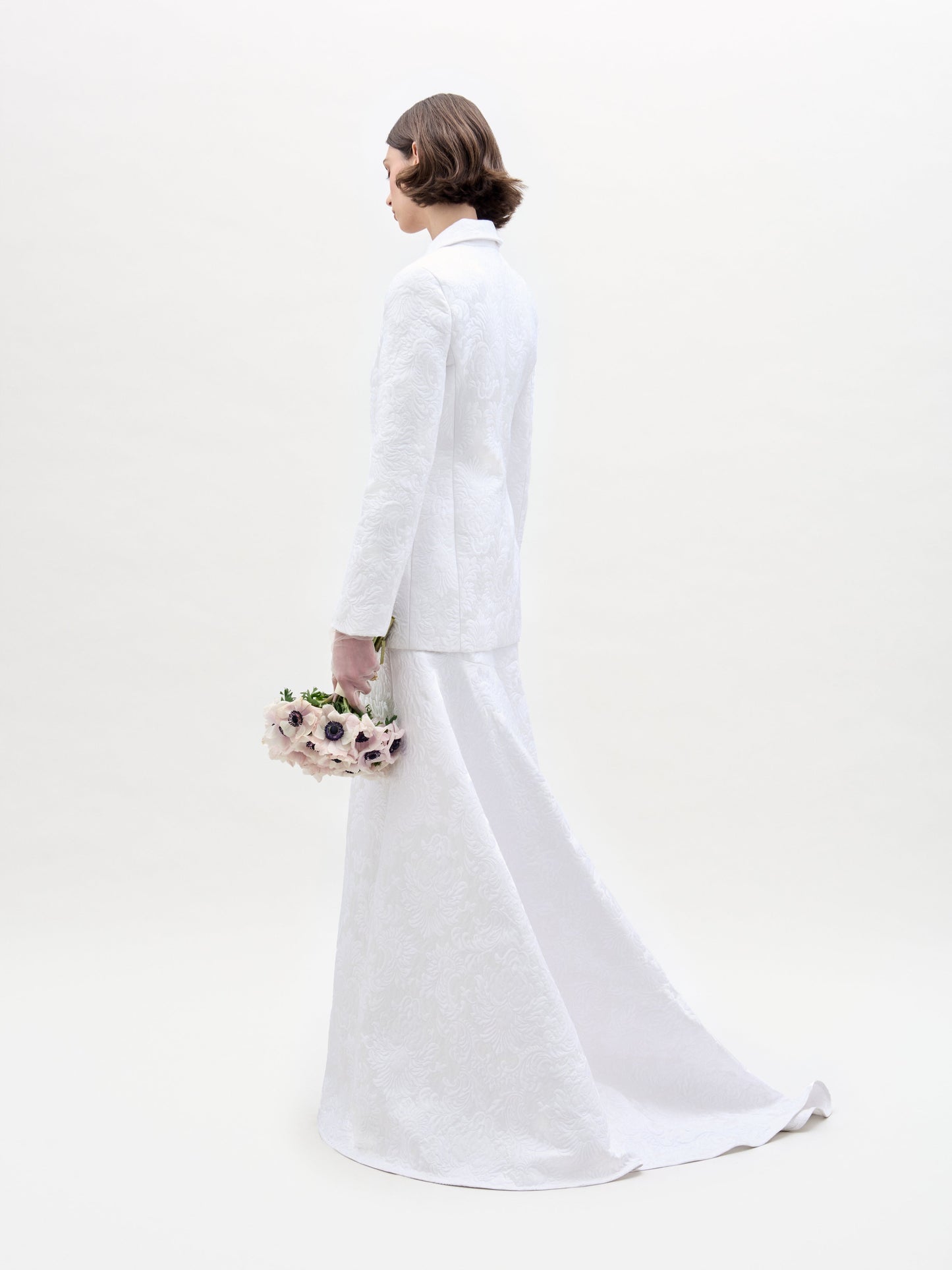 A person with short brown hair wears the Imola Skirt— a long, high-waisted white maxi skirt with a matching Italian fabric jacket—holding pale flowers and standing against a plain light background.