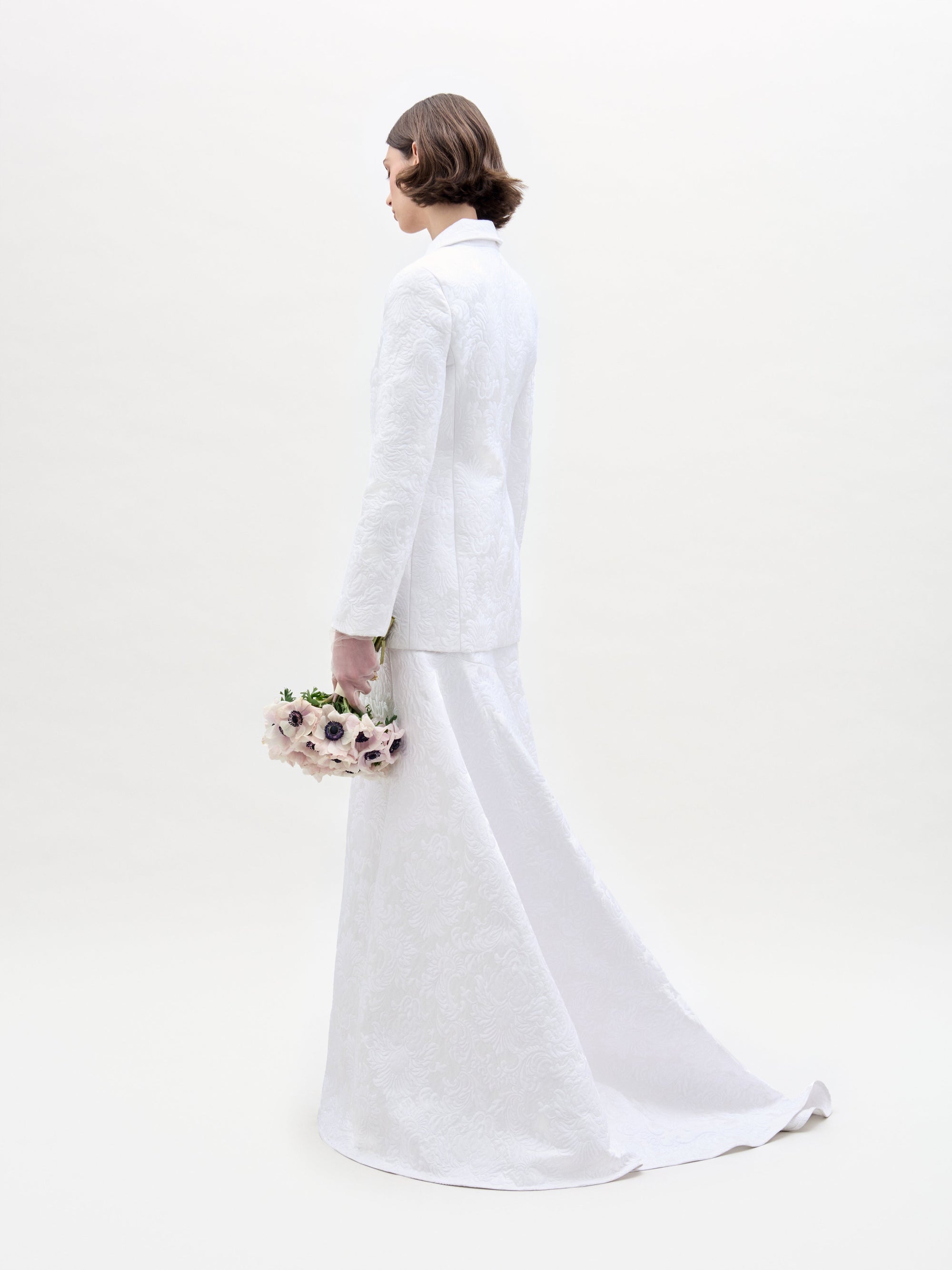 A person with short brown hair wears the Imola Skirt— a long, high-waisted white maxi skirt with a matching Italian fabric jacket—holding pale flowers and standing against a plain light background.