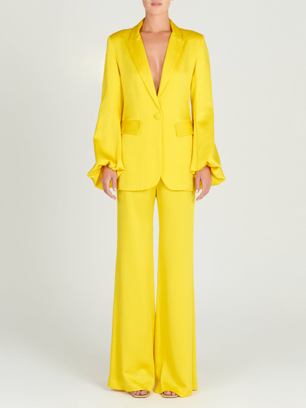 A person in the Coco Blazer—a bright yellow hammered satin jacket with flared sleeves and flap pockets—styled with wide-leg trousers and open-toe heels for a chic, softly tailored look.