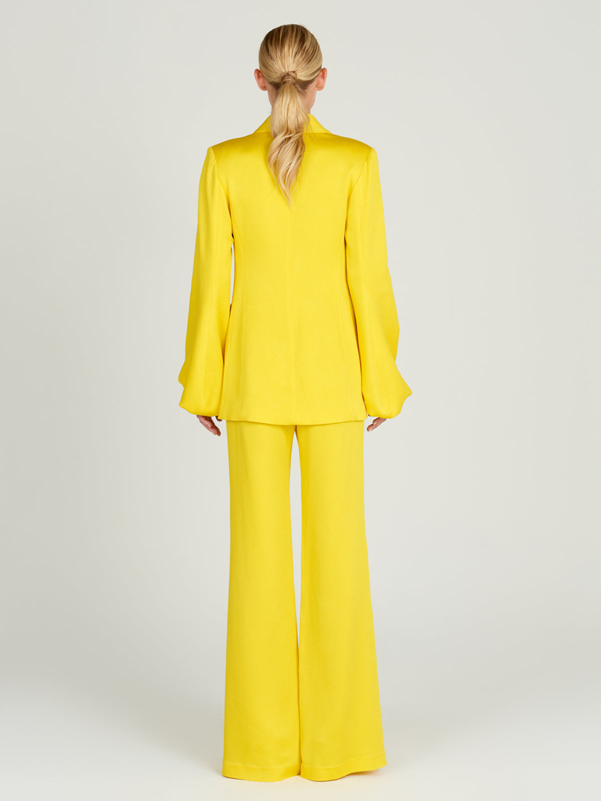 A woman with blonde hair in a low ponytail faces away from the camera, wearing the bright yellow Coco Blazer and wide-legged pants, highlighting soft tailoring against a plain, light backdrop.