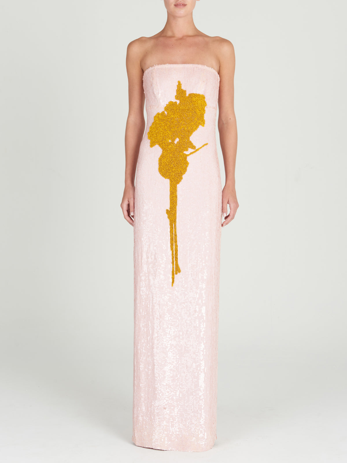 Someone wears the Margaret Dress, a pale pink strapless column gown with a large gold sequin abstract design on the front, standing against a plain light background.