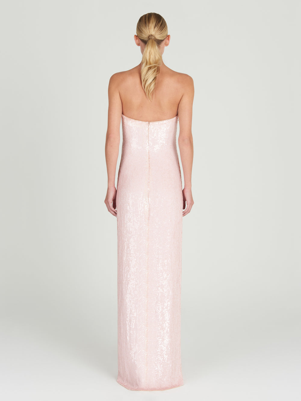 A woman with blonde hair in a low ponytail is seen from behind wearing the Margaret Dress, a floor-length, strapless column gown in pale pink with sequin embroidery. The background is plain and light-colored.