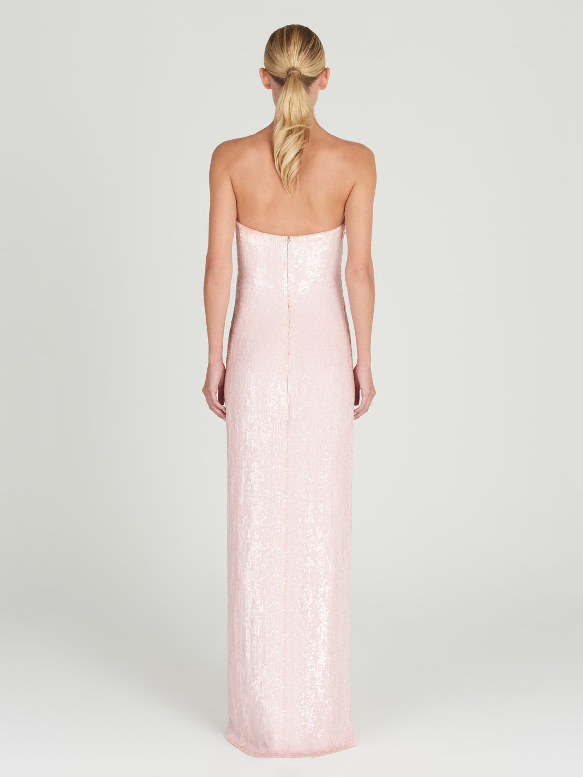 A woman with blonde hair in a low ponytail is seen from behind wearing the Margaret Dress, a floor-length, strapless column gown in pale pink with sequin embroidery. The background is plain and light-colored.