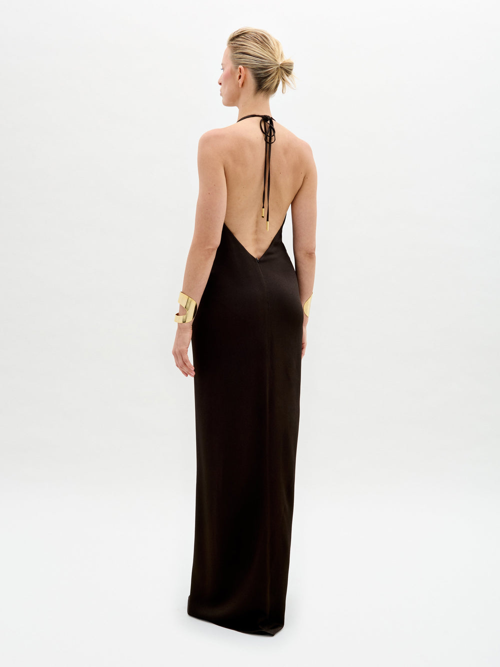A person stands against a white background in the Rowan Dress, featuring a sleeveless deep V-neck black hammered satin maxi silhouette with a central gold embellishment, paired with gold bracelets, short hair, and gold high-heeled shoes.