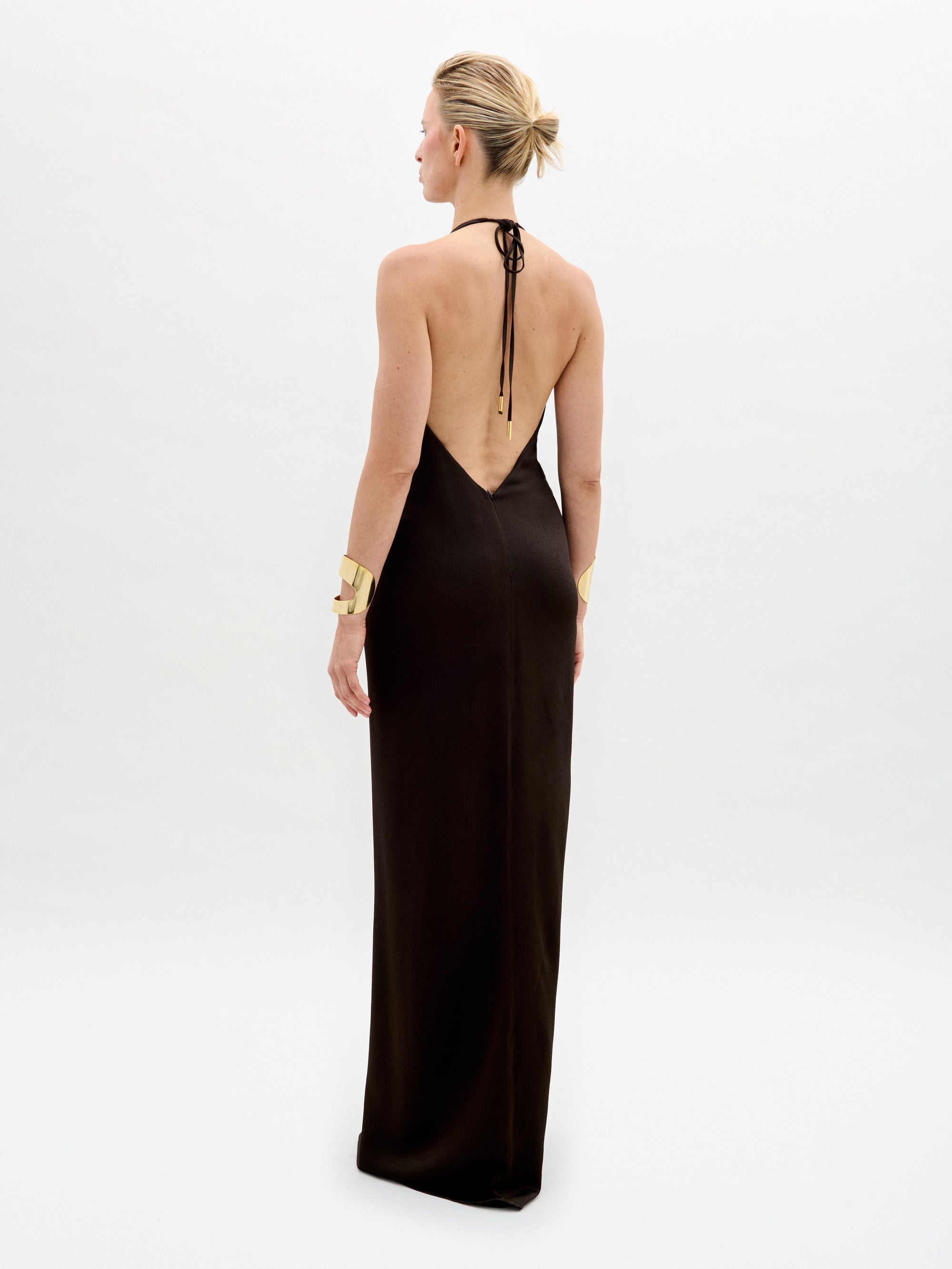 A person stands against a white background in the Rowan Dress, featuring a sleeveless deep V-neck black hammered satin maxi silhouette with a central gold embellishment, paired with gold bracelets, short hair, and gold high-heeled shoes.