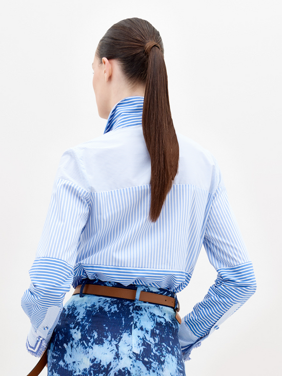 A person stands wearing a Raina Shirt Multi Blue Stripes, blue patterned pants, and a brown belt with a silver buckle. The background is plain white. Pre-order now to ensure it ships by the November 15th, 2024 date.