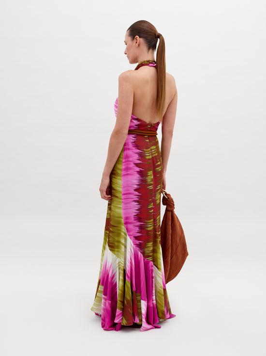 A person dressed in a Raisa Dress Violeta Lime Tie Dye with pink, green, and white patterns, holding a large brown bag, posing against a plain background. Pre-order by November 15th 2024 to ensure the perfect ship-by date.