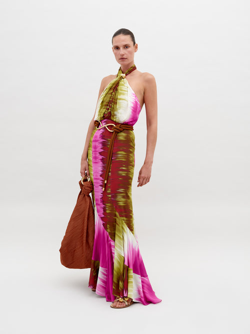 A person dressed in a Raisa Dress Violeta Lime Tie Dye with pink, green, and white patterns, holding a large brown bag, posing against a plain background. Pre-order by November 15th 2024 to ensure the perfect ship-by date.