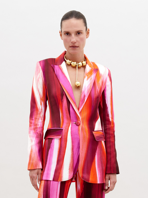 Woman wearing a vibrant, multi-colored striped Rebeca Jacket Magenta Abstract Waves with a deep neckline, accessorized with a chunky gold choker necklace, standing against a plain white background. Pre-order now and have it ship by date of November 15th, 2024.