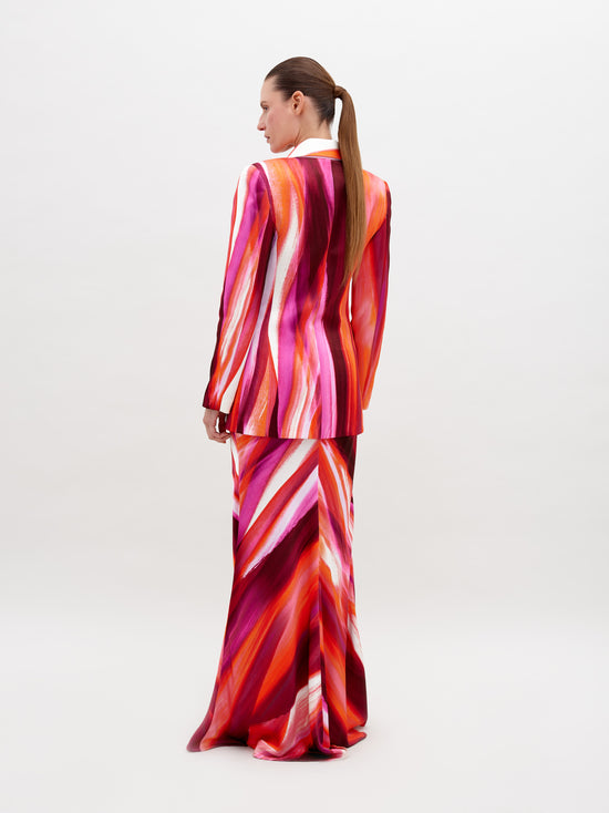 A person stands against a plain background, wearing a vibrant suit in red, pink, and orange streaks, paired with the Laurina Skirt Magenta Abstract Waves and bold gold necklace. This stunning ensemble will be available for pre-order soon and is set to ship by date 2024.