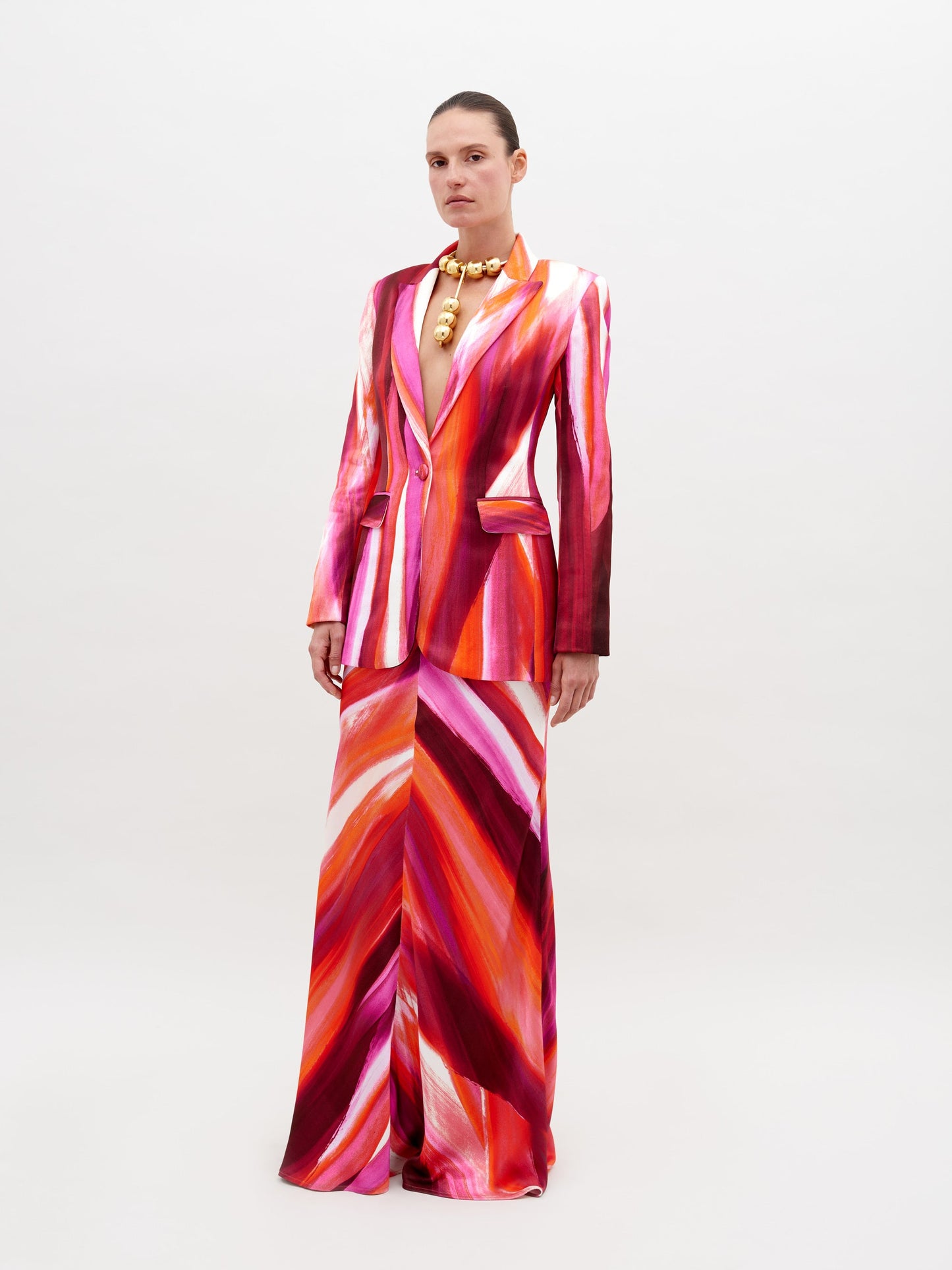 A person stands against a plain background, wearing a vibrant suit in red, pink, and orange streaks, paired with the Laurina Skirt Magenta Abstract Waves and bold gold necklace. This stunning ensemble will be available for pre-order soon and is set to ship by date 2024.