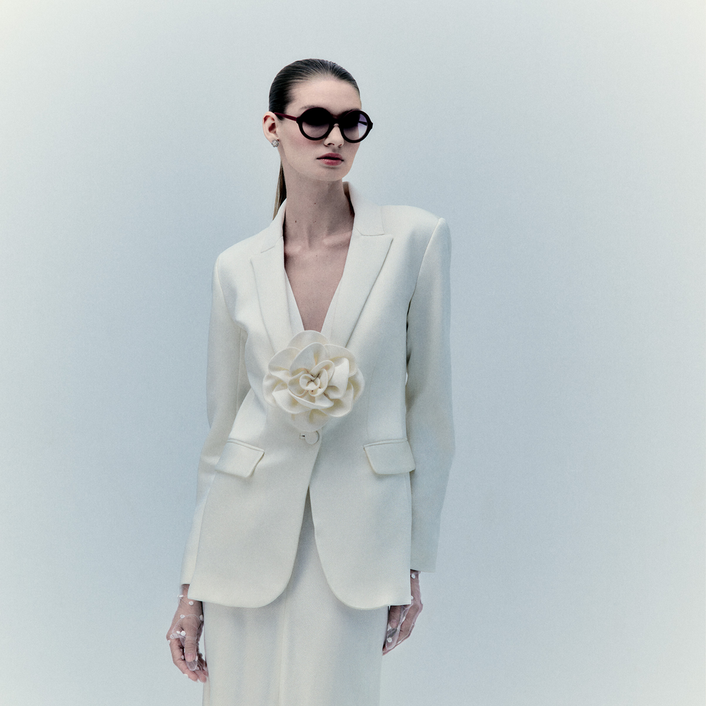 A woman in the Daniella Dress, a cream suit featuring a bold 3D floral appliqué on the lapel, stands against a neutral background. She wears dark sunglasses and has slicked-back hair for a modern, elegant style.