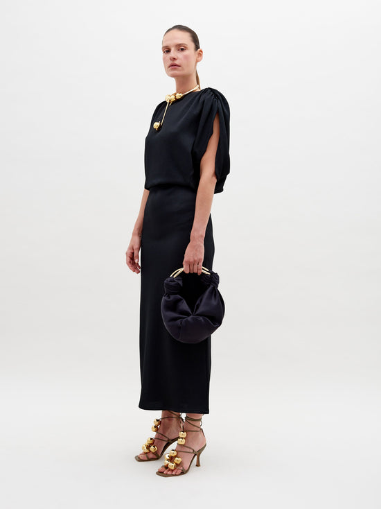 Against a plain background, a person stands elegantly in the Reese Dress Black with gold accessories, holding a matching black handbag. They also sport high-heeled sandals adorned with gold embellishments. Pre-order now to ensure your look is ready by the ship-by date of November 15th, 2024.