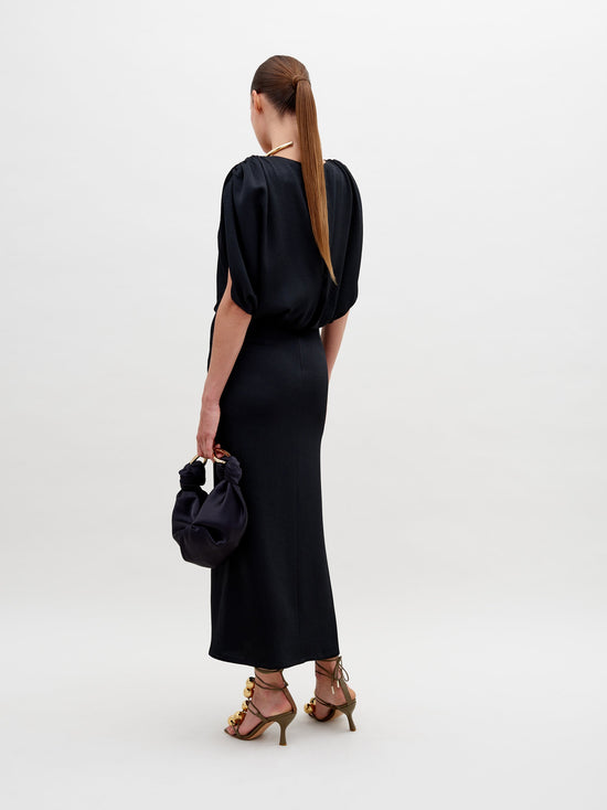 Against a plain background, a person stands elegantly in the Reese Dress Black with gold accessories, holding a matching black handbag. They also sport high-heeled sandals adorned with gold embellishments. Pre-order now to ensure your look is ready by the ship-by date of November 15th, 2024.