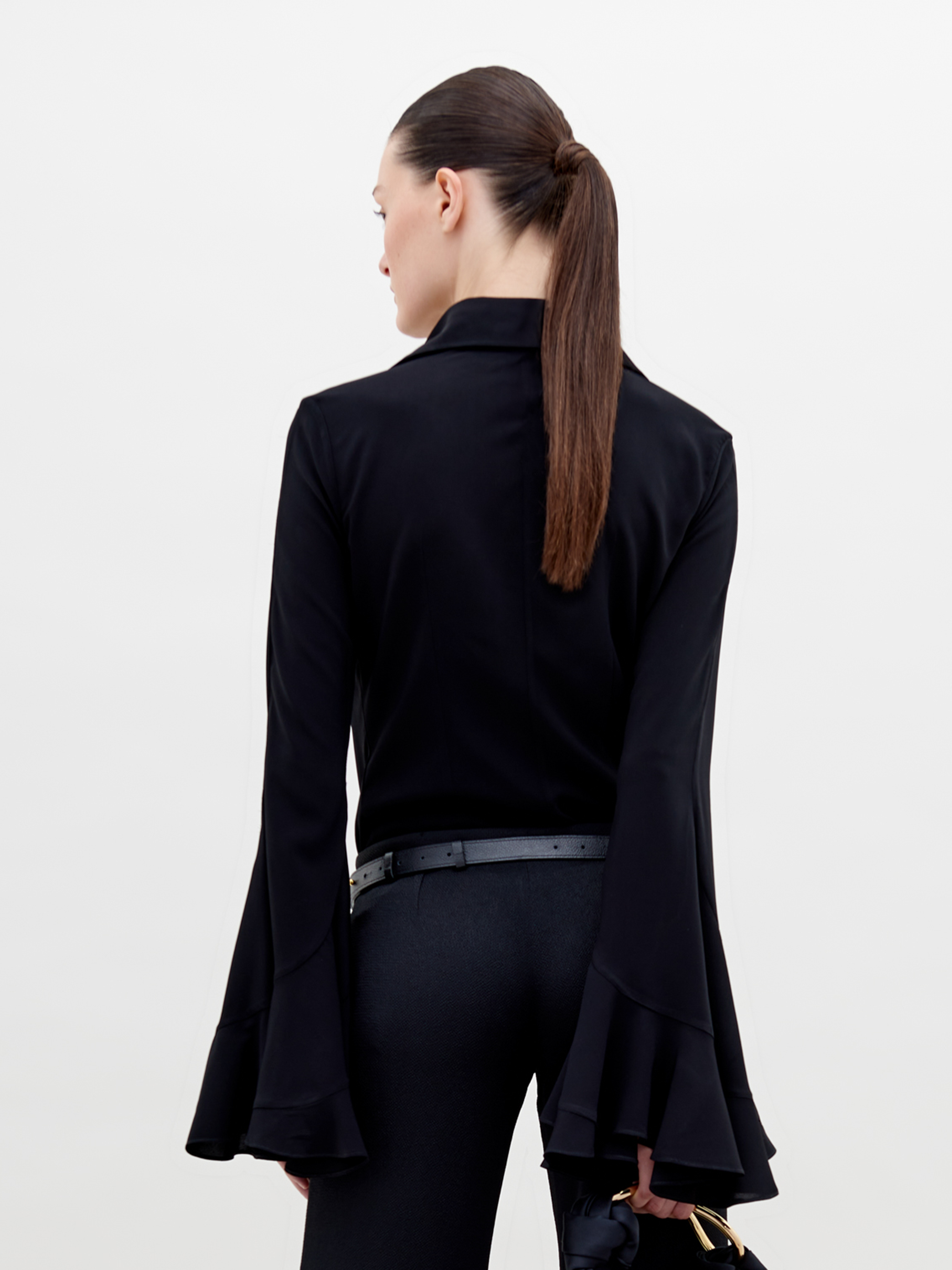 A person with slicked-back hair wears a black outfit with flared sleeves, a gold necklace, and a belt featuring a large gold infinity symbol. They hold the Reggie Blouse Black in their hand, available for pre-order now and set to ship by date November 15th, 2024.