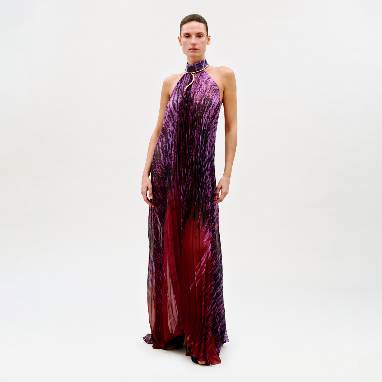 A person stands against a light background, wearing the Lucia Dress—a floor-length halter-neck gown featuring a purple and red abstract pattern with a metallic ring detail at the neckline.