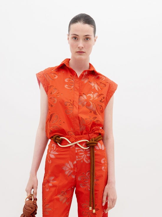 A person stands wearing a vibrant orange sleeveless top and matching pants with floral patterns, accessorized with a brown belt and holding a brown handbag. Pre-order the Rina Blouse Rouge Embroidery now to ensure it ships by the date of November 15th, 2024.