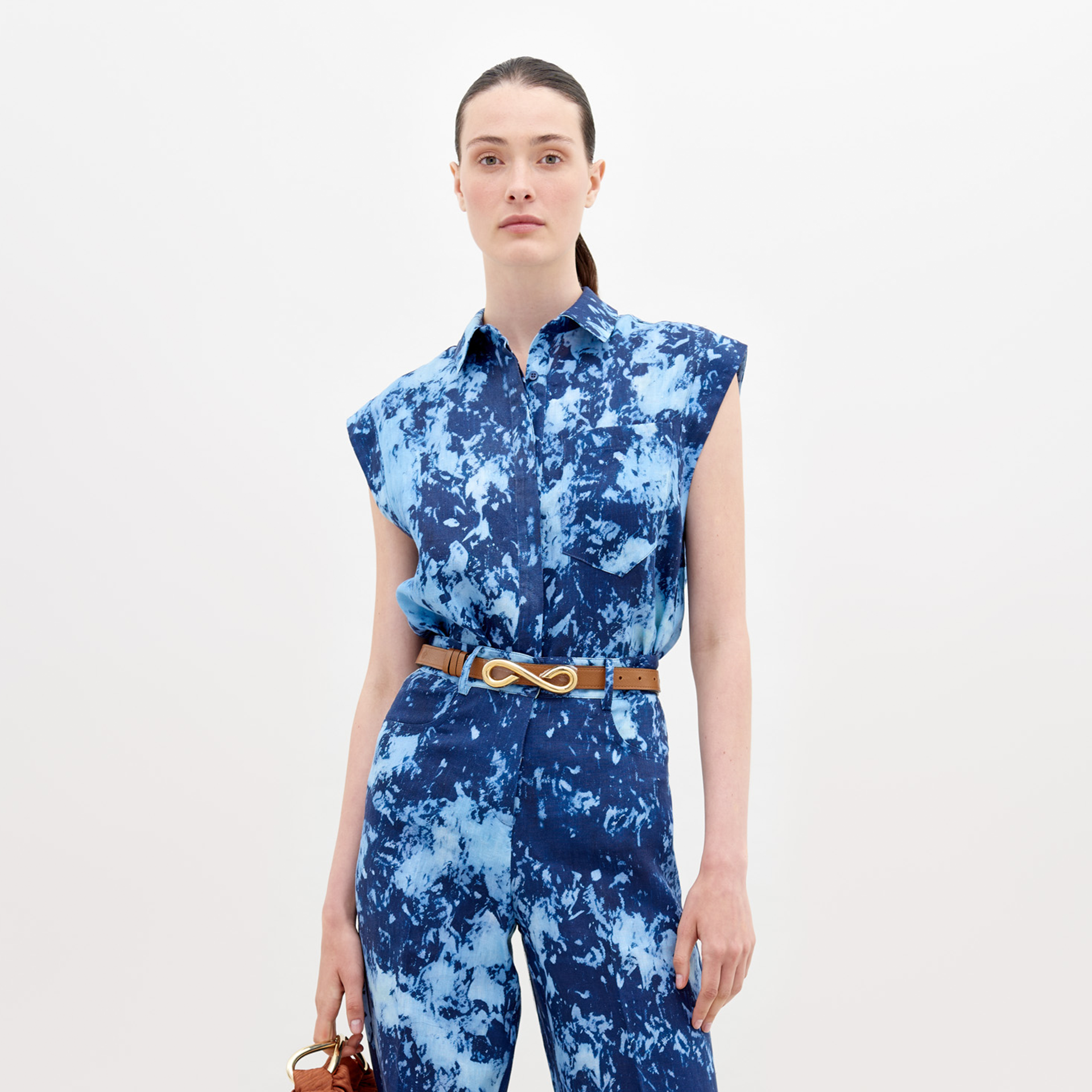 A woman wearing a blue tie-dye dress with a collar and sleeveless design, carrying a large navy blue bag on her shoulder, with her hair tied back in a ponytail, is showcasing the Rina Blouse Indigo exclusive pre-order item available until November 15th, 2024.