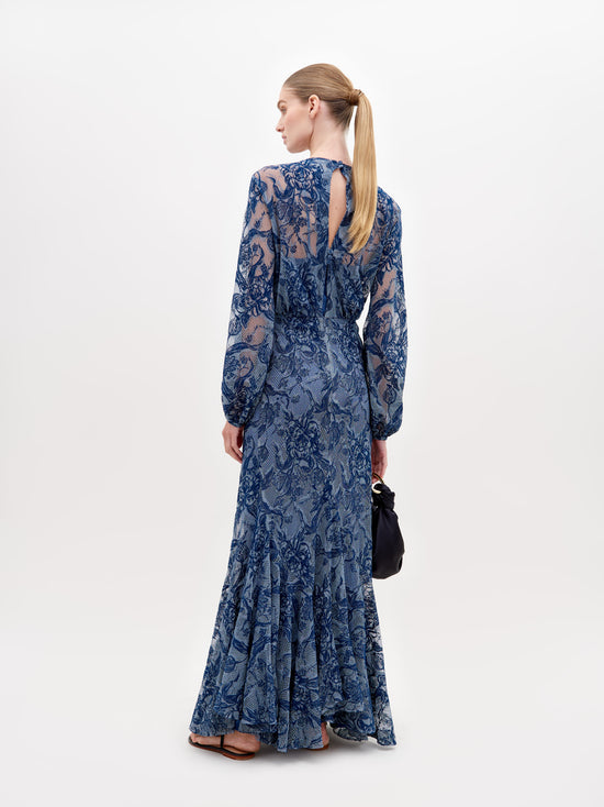 A person in a long, blue lace dress with a deep V-neckline and long sleeves stands against a plain white background, holding a black clutch—the Robin Dress Indigo Lace, available for pre-order, set to ship by November 15th, 2024.