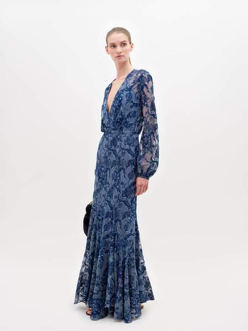 A person in a long, blue lace dress with a deep V-neckline and long sleeves stands against a plain white background, holding a black clutch—the Robin Dress Indigo Lace, available for pre-order, set to ship by November 15th, 2024.