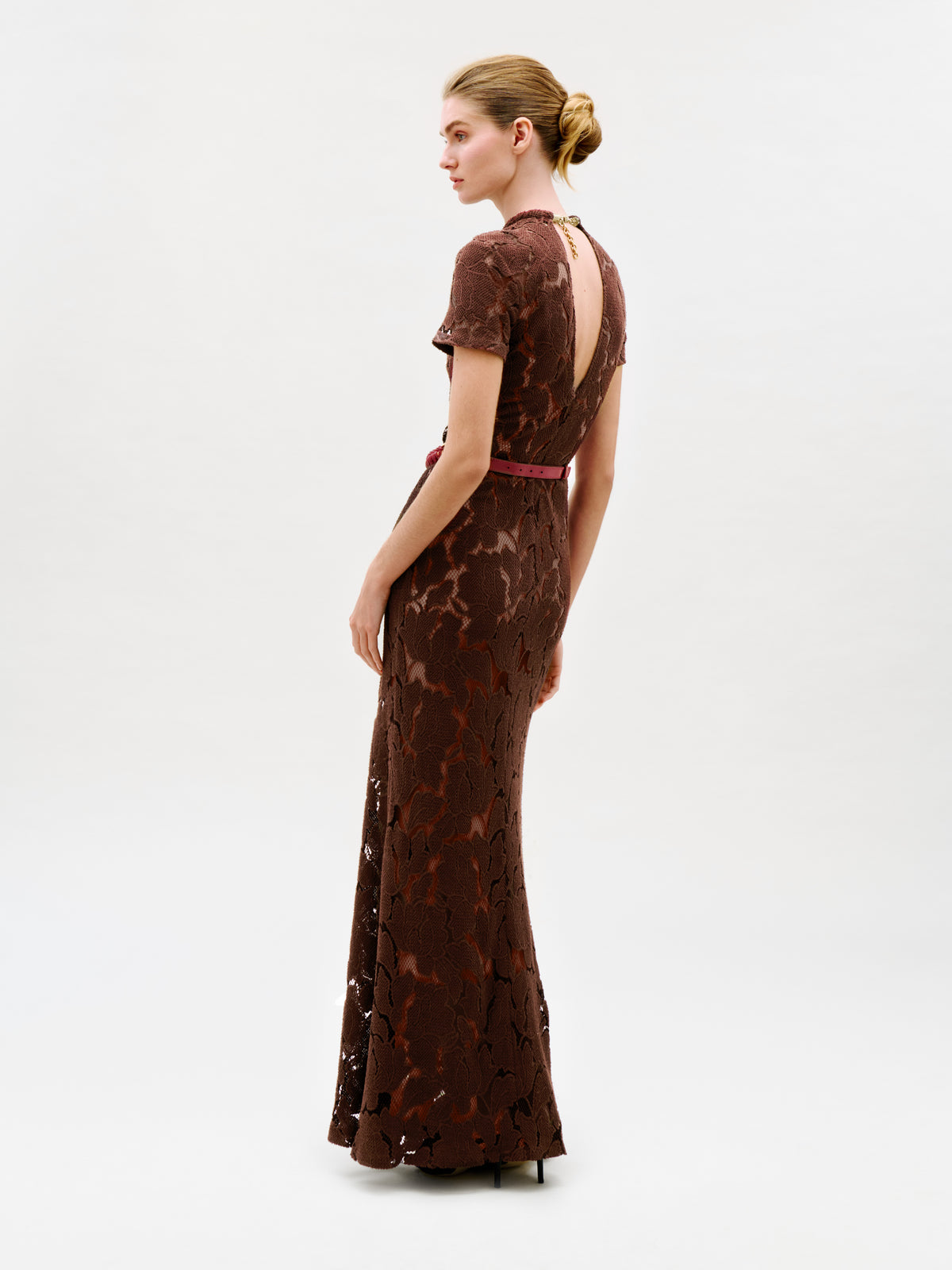 A woman stands sideways against a plain light background, looking to her left, wearing the Rochelle Dress—a long brown lace maxi dress with short sleeves and an open back. Crafted from luxurious Italian fabric and made in Colombia, she wears her hair in a neat bun and pairs the look with high heels.