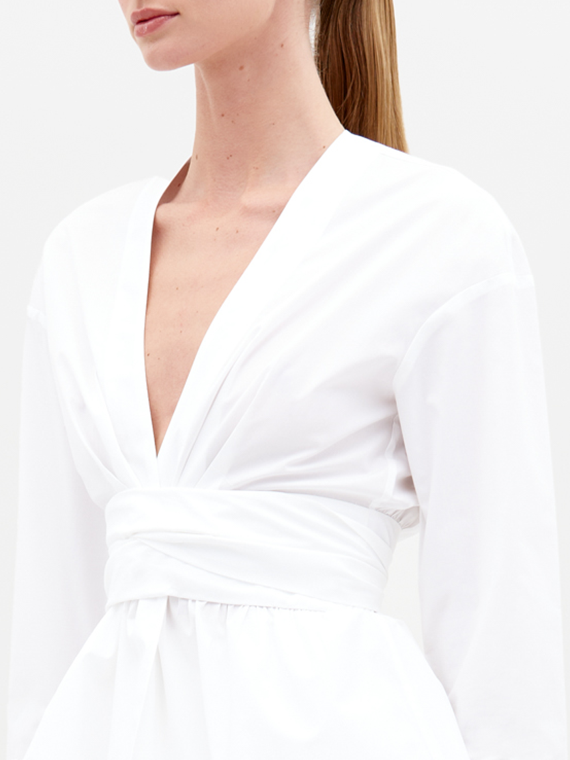 A woman with light skin and straight, tied-back hair wears the Saanvi Blouse in white Italian fabric, featuring a deep V-neck, long sleeves, and a wide ruched waistband—shown from shoulders to waist against a plain background.