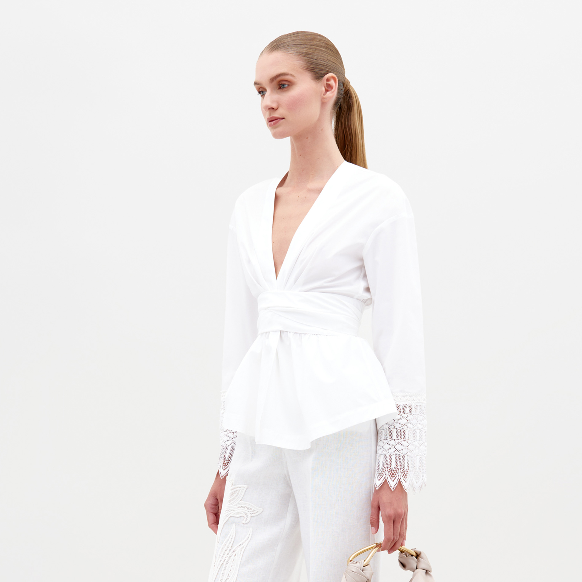 A woman with straight, pulled-back hair wears the Saanvi Blouse—a white cotton top featuring a wrap waist tie and lace cuffs—paired with white embroidered trousers and a pale handbag, standing against a plain white background.