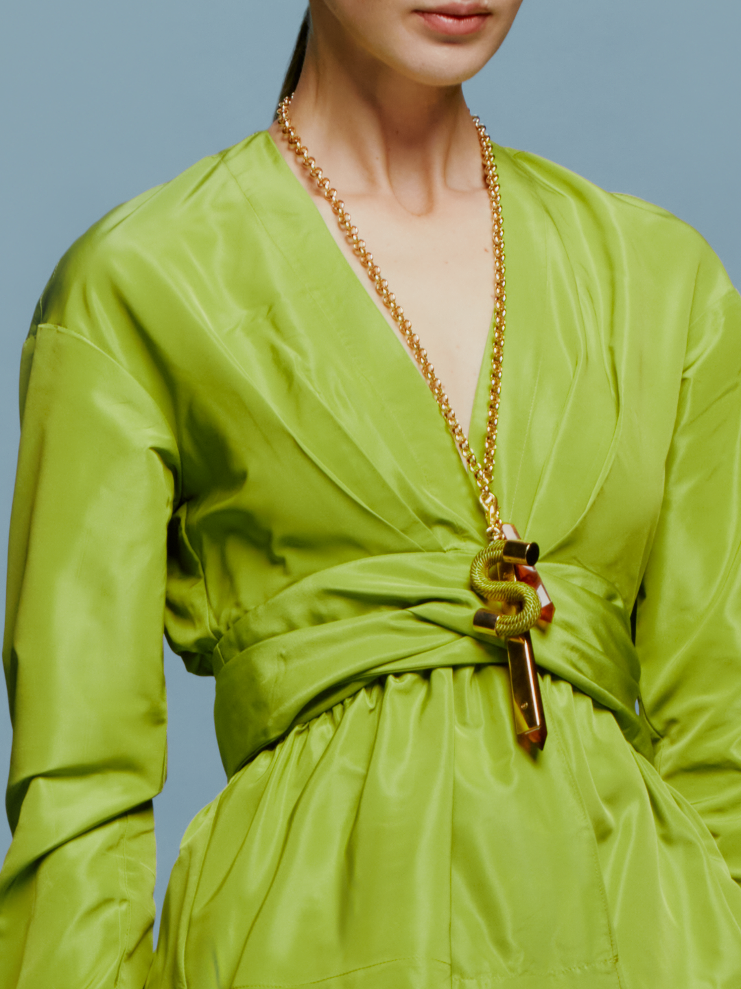 Wearing the elegant Saanvi Blouse in Lime, paired with a gold necklace and matching handbag, a woman stands against a plain blue background, exuding effortless style as she embraces the calm of September 2nd.