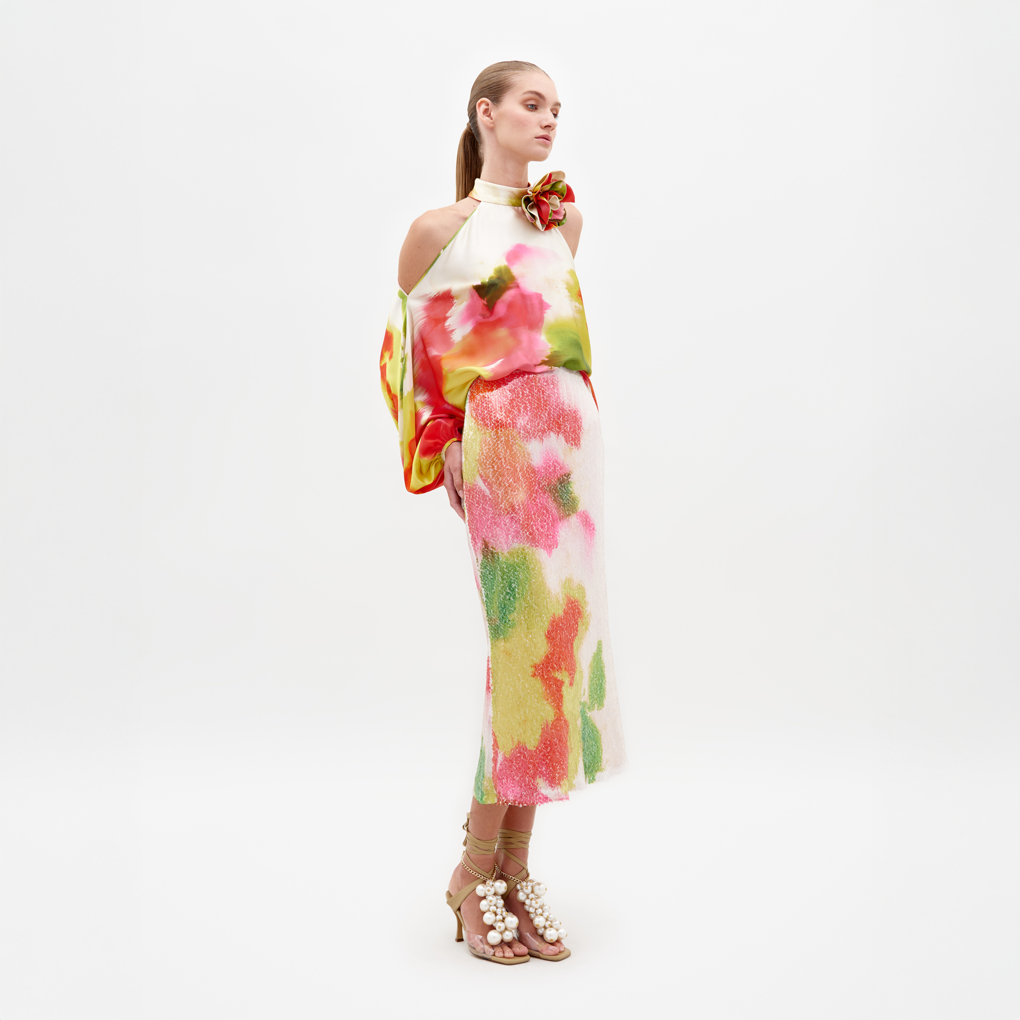 A person stands facing slightly to the side, wearing a colorful off-shoulder top and the Sarla Skirt Spring Blooms midi skirt, paired with high heels adorned with large pearl decorations; pre-order now to ensure it ships by February 15th, 2025.