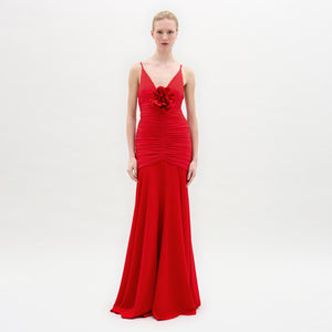 A woman poses against a white background in the Sana Dress—a fitted red maxi made from Italian fabric, with thin straps, 3D floral appliqué at the chest, a ruched bodice, and a beautifully flared skirt.