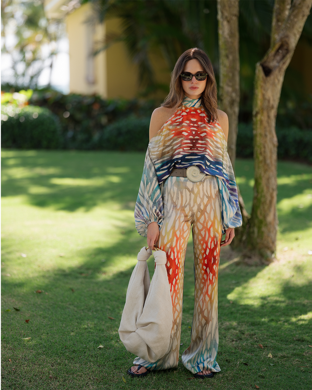A woman stands confidently in a vibrant, patterned outfit with tones of orange, blue, and beige. She holds a textured bag. The backdrop is a sunny garden with lush greenery, evoking a relaxed yet stylish vibe.