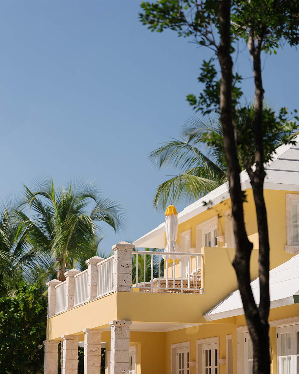 Yellow two-story house with white railings and a sun umbrella on the balcony. Surrounded by palm trees under a clear blue sky, creating a tropical feel.