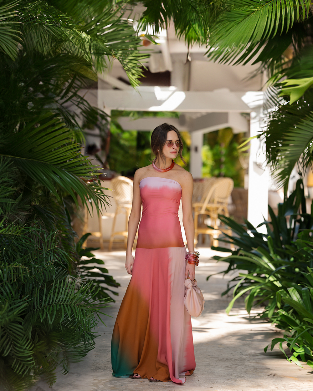 Sofia Tcherassi in a flowing pink and brown ombre dress stands on a sunlit pathway surrounded by lush green tropical plants, exuding a relaxed and stylish vibe.
