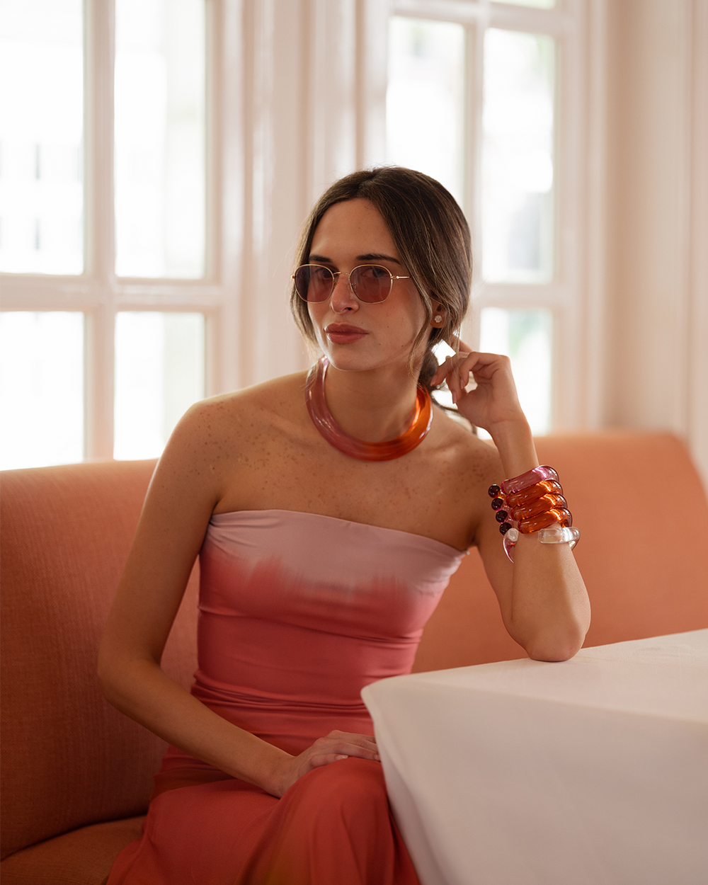 A woman in a strapless coral dress sits by a window on a peach sofa. She wears orange jewelry, sunglasses, and has a relaxed, elegant expression.