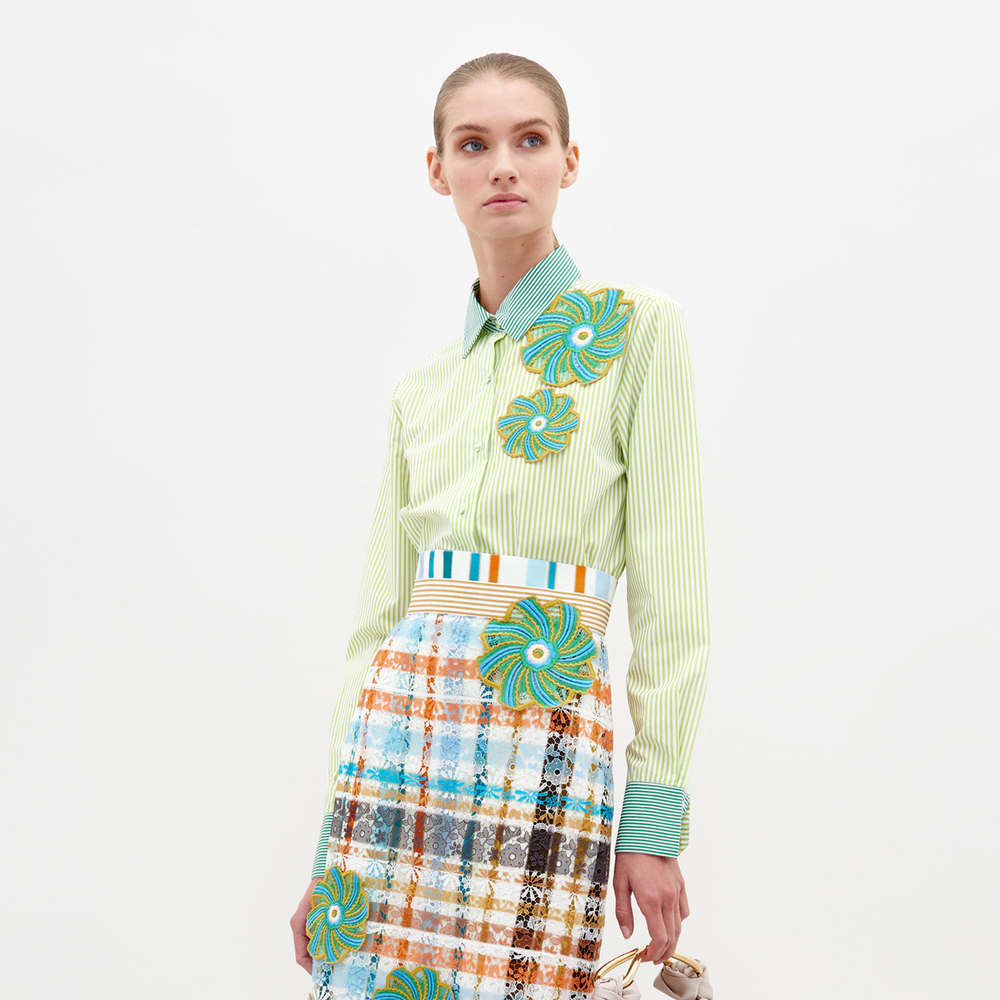 A woman stands against a plain background wearing the Shao Blouse Green Stripes, adorned with colorful floral embellishments, and a multicolored plaid skirt. She holds a small item in her left hand, now available for pre-order and expected to ship by February 15th, 2025.