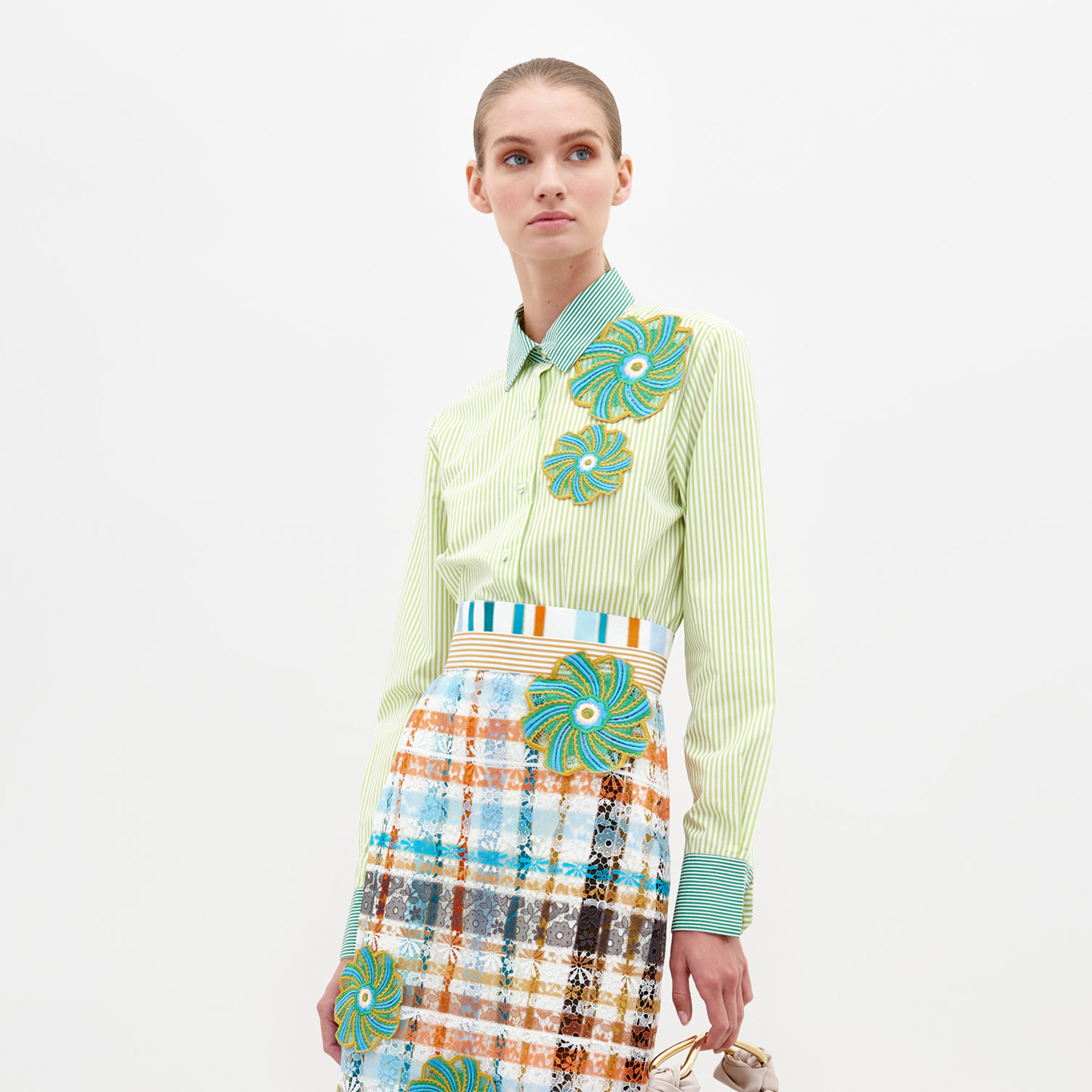 A woman stands against a plain background wearing the Shao Blouse Green Stripes, adorned with colorful floral embellishments, and a multicolored plaid skirt. She holds a small item in her left hand, now available for pre-order and expected to ship by February 15th, 2025.
