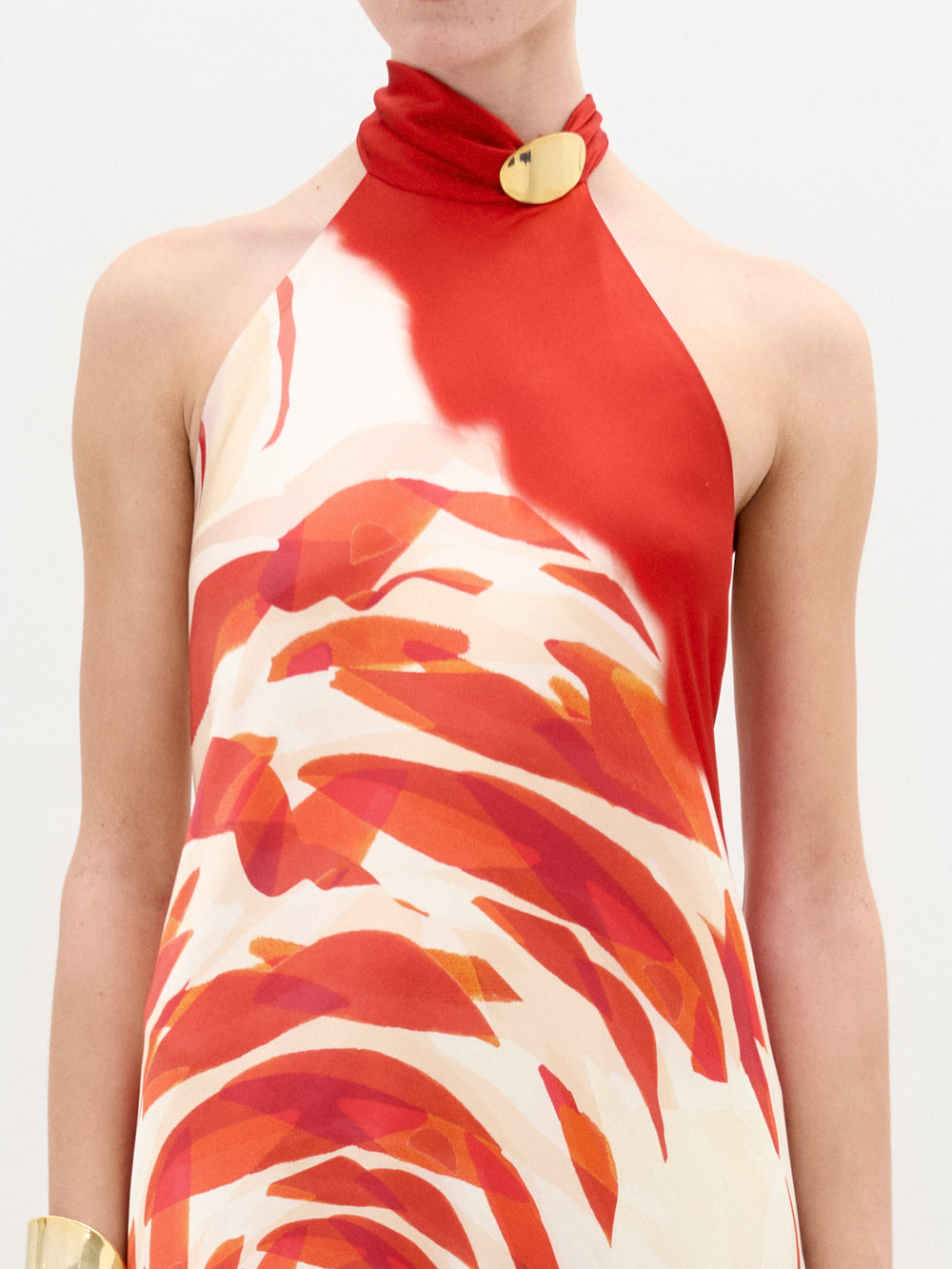 A person stands against a white background wearing the Sherry Dress—a red and white silk maxi with a bold swirling rose pattern, halter neckline, draped column fit—styled with gold accessories.