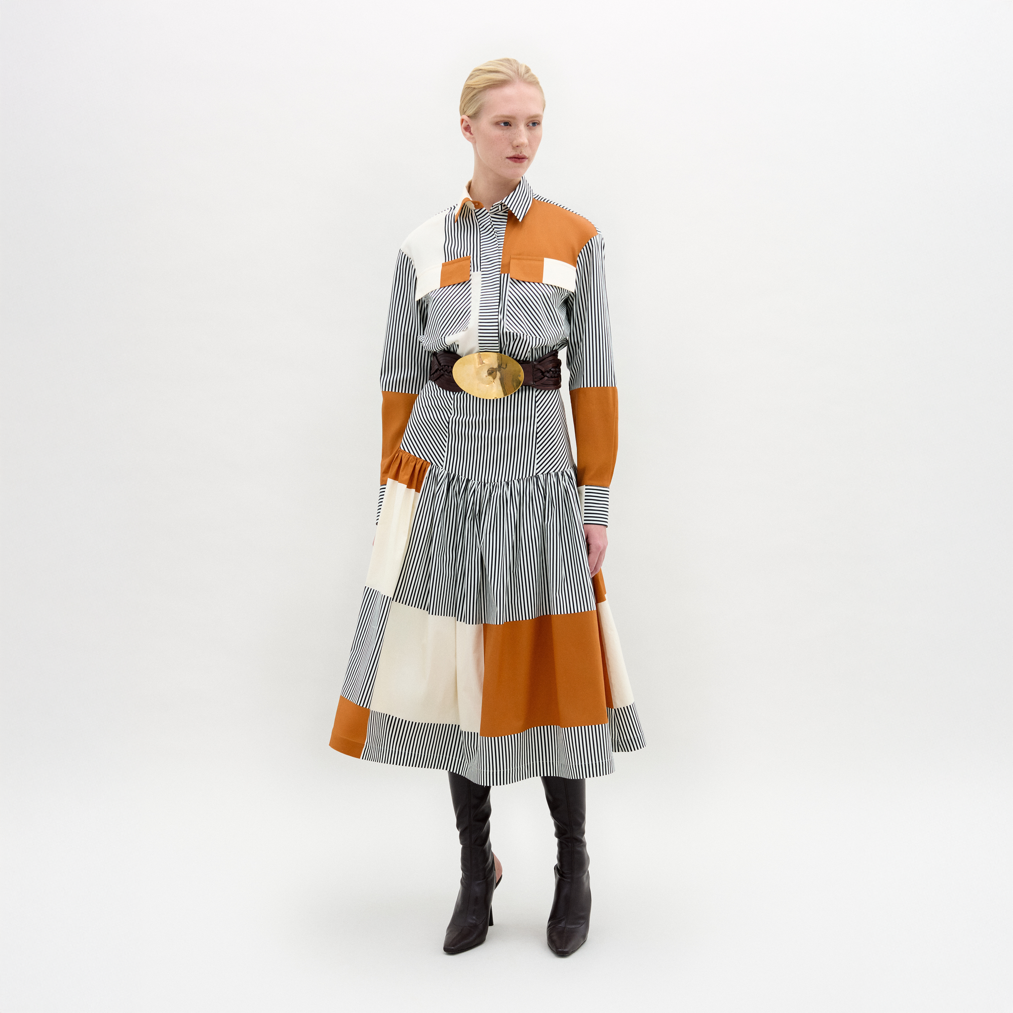 A woman stands against a white background wearing the Shoreh Skirt, a patchwork design in Italian fabric with vertical stripes and orange, white, and brown blocks, styled with a wide belt featuring a large gold buckle and knee-high black boots.