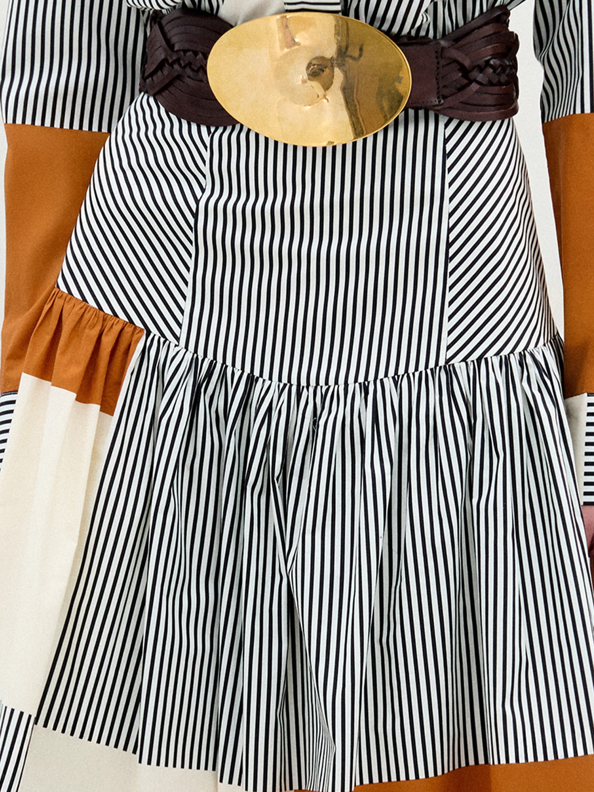 A close-up of a person wearing the Shoreh Skirt, a black-and-white vertical striped pleated midi skirt with panels of solid brown and white Italian fabric, styled with a wide brown belt featuring a large round gold buckle.