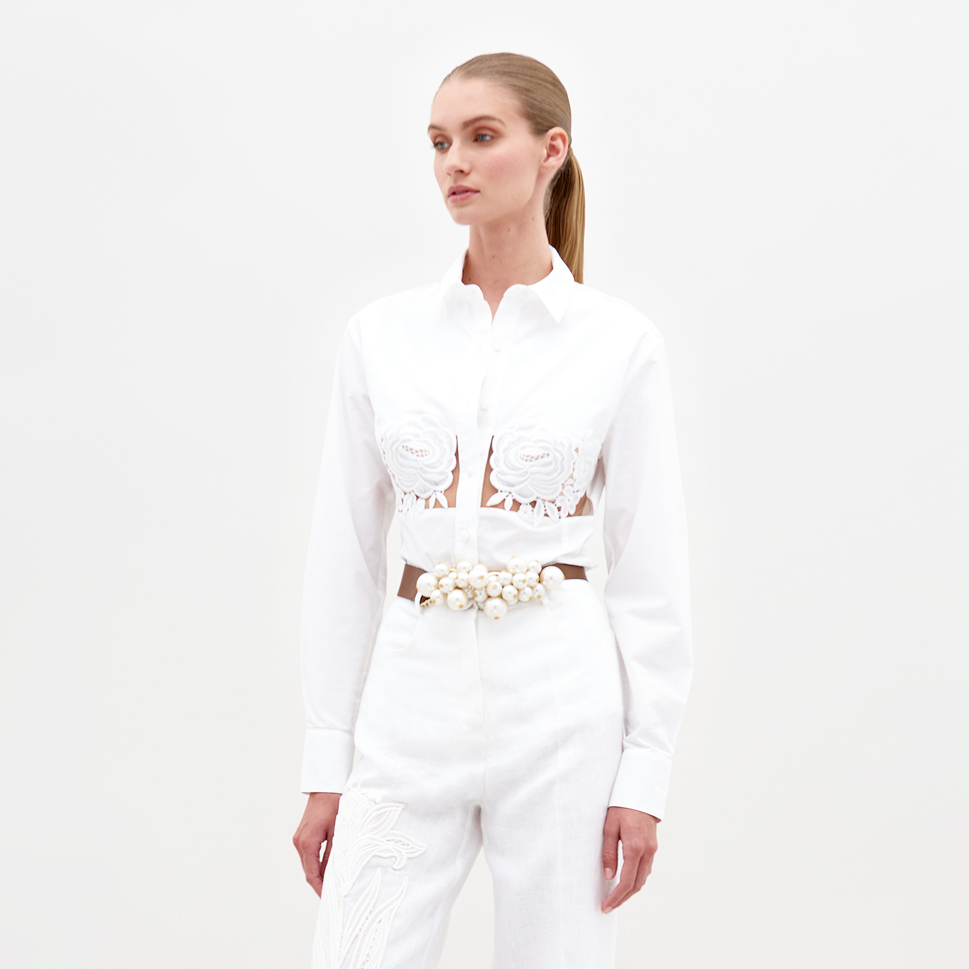 A woman with long, slicked-back hair wears the Sigrid Blouse—white cotton with chest cutouts—paired with high-waisted floral-embellished pants and a pearl belt, standing against a plain white background.